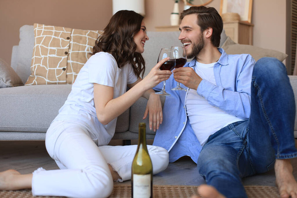 Happy couple sitting, relaxing on floor in living room, drinking red wine. Smiling young husband and wife rest at home enjoy romantic date on family weekend together. - Fotografie, Obrázek