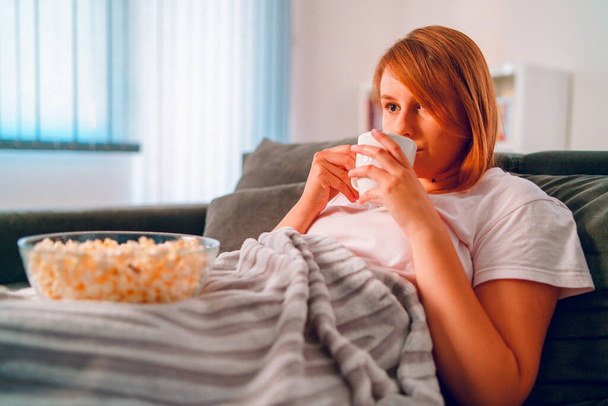 Νεαρή γυναίκα που κάθεται στον καναπέ του κρεβατιού στο σπίτι, ξαπλωμένη με ένα μπολ ποπ κορν βλέποντας τηλεόραση καλυμμένη με κουβέρτα στο διαμέρισμά της μόνη της να απολαμβάνει ταινίες ή σειρές πίνοντας ένα φλιτζάνι τσάι ή καφέ - Φωτογραφία, εικόνα