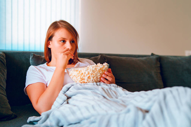 Νεαρή γυναίκα που κάθεται στον καναπέ του κρεβατιού στο σπίτι, ξαπλωμένη με ένα μπολ ποπ κορν βλέποντας τηλεόραση καλυμμένη με κουβέρτα στο διαμέρισμά της μόνη της να απολαμβάνει ταινίες ή σειρές - Φωτογραφία, εικόνα