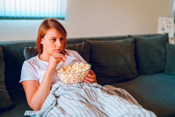 Young woman sitting on the bed sofa at home, lying with a popcorn bowl watching TV covered with blanket at her apartment alone enjoying movies or series - 写真・画像