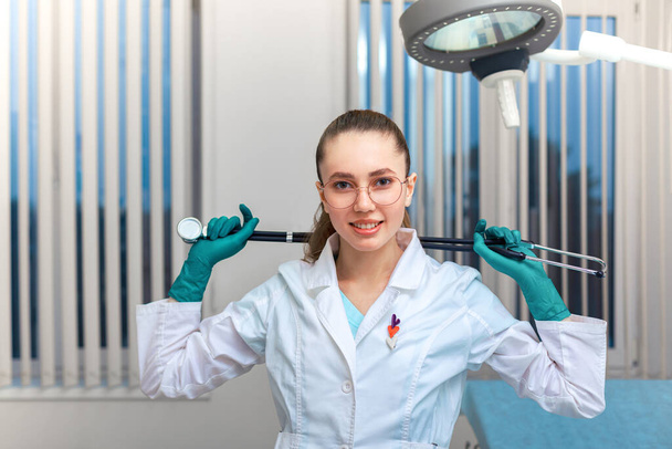 Mladá doktorka v kabátě s fonendoskopem v rukou pózující na kameru na pozadí oddělení v nemocnici. Kopírovat prostor, Koncept medicíny - Fotografie, Obrázek