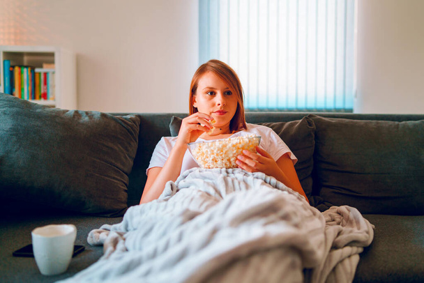 Młoda kobieta siedząca na kanapie w domu, leżąca z misą popcornu i oglądająca telewizję pokrytą kocem w swoim mieszkaniu, ciesząca się filmami lub serialami - Zdjęcie, obraz