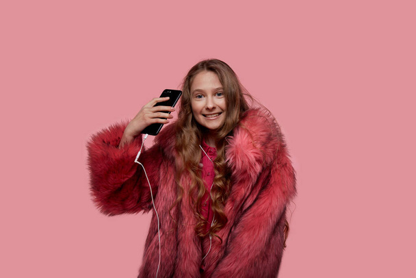 girl turns on songs on the phone and looks at the camera in a pink fur coat - Photo, Image