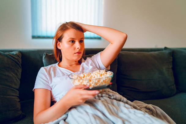 Młoda kobieta siedząca na kanapie w domu, leżąca z misą popcornu i oglądająca telewizję pokrytą kocem w swoim mieszkaniu, ciesząca się filmami lub serialami - Zdjęcie, obraz