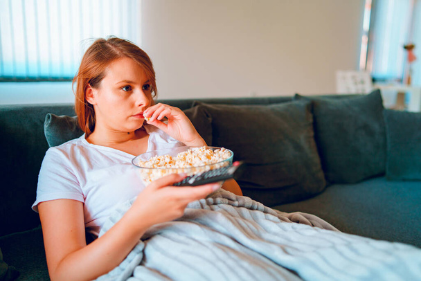 Νεαρή γυναίκα που κάθεται στον καναπέ του κρεβατιού στο σπίτι, ξαπλωμένη με ένα μπολ ποπ κορν βλέποντας τηλεόραση καλυμμένη με κουβέρτα στο διαμέρισμά της μόνη της να απολαμβάνει ταινίες ή σειρές - Φωτογραφία, εικόνα