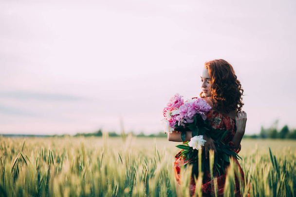 Red-haired girl in a red dress with a bouquet of peonies flowers in a summer wheat field at sunset. Soft focus. Bouquet for the holiday. - Photo, image