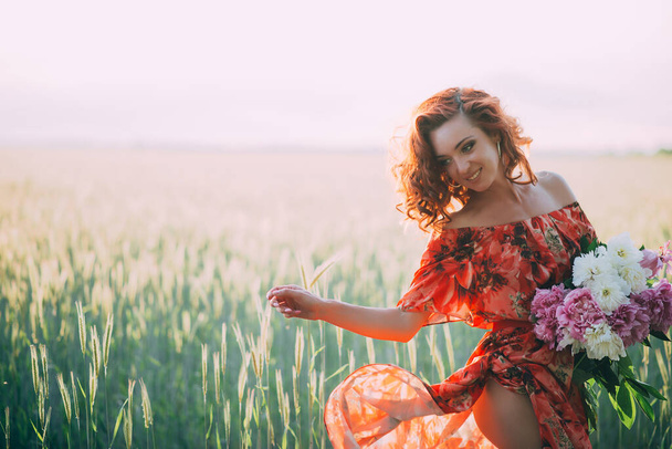 Zrzavá dívka v červených šatech s kyticí pivoňek tančících radostně na pšeničném poli v létě při západu slunce. Jemné soustředění. Zadní světlo. - Fotografie, Obrázek