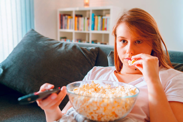 Young woman sitting on the bed sofa at home, lying with a popcorn bowl watching TV covered with blanket at her apartment alone enjoying movies or series holding remote control pointed - Photo, image