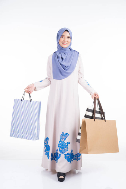 Ένα όμορφο μουσουλμανικό θηλυκό μοντέλο σε ένα ασιατικό παραδοσιακό φόρεμα jubah μεταφέρουν τσάντες ψώνια που απομονώνονται σε λευκό φόντο. Eidul fitri εορταστική έννοια ψώνια. Πλήρες μήκος πορτρέτο. - Φωτογραφία, εικόνα