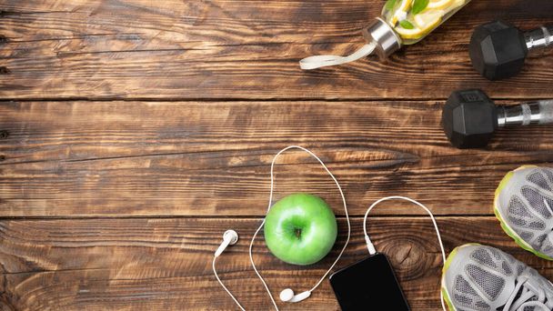Fitness equipment on wooden background with copy space: dumbbells, phone, headphones, green apple, sneakers. Healthy lifestyle concept, template for recipes, workouts, reusable products, zero waste - Photo, Image