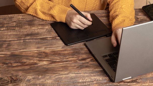 Kobieta pracująca na tablecie graficznym przy stole, laptop w biurze. Koncepcja zdalnej pracy w domu podczas kwarantanny z powodu pandemii. Zostań w domu. Kobiece ręce i żółty sweter - Zdjęcie, obraz