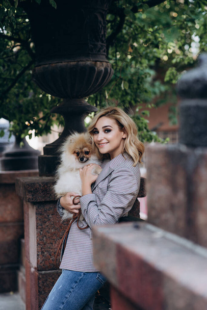 Beautiful young blond woman with her pet Pomeranian Spitz dog breed for a walk. Little fluffy dog. Soft selective focus. - Фото, изображение
