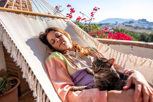 Genç İspanyol kadın beyaz hamakta tekir kedisiyle birlikte evdeki terasta yatıyor. - Fotoğraf, Görsel