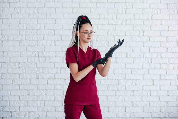 a girl with African pigtails on her head in a red medical uniform wears black gloves. she stands against a white brick wall - Foto, Imagen