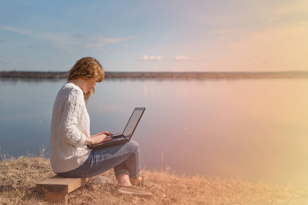 woman working remotely on a laptop outdoors, on a background of river and sky - Φωτογραφία, εικόνα