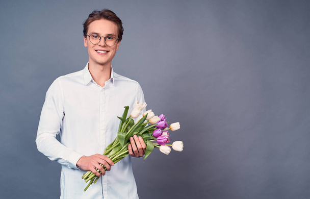 Delicate tulips for her. A young man with blond hair and blue eyes, in a white shirt and round glasses with a bouquet of tulips. Handsome man holds pink and white tulips. Copy space. - Foto, Bild