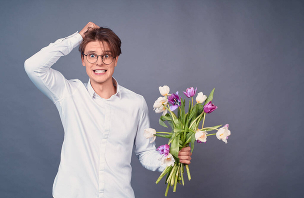 Puzzled, bewildered young man holds a large bouquet of broken white and pink tulips, his hairstyle is spoiled, an unsuccessful date, wears a white shirt, isolated on a gray background. Copy space. - 写真・画像
