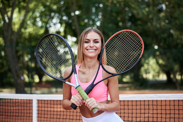 Young blond woman, wearing pink top and white skirt, holding two tennis rackets on court with orange ground in summer. Three-quarter portrait of sportswoman, training outside. Active healthy life. - 写真・画像