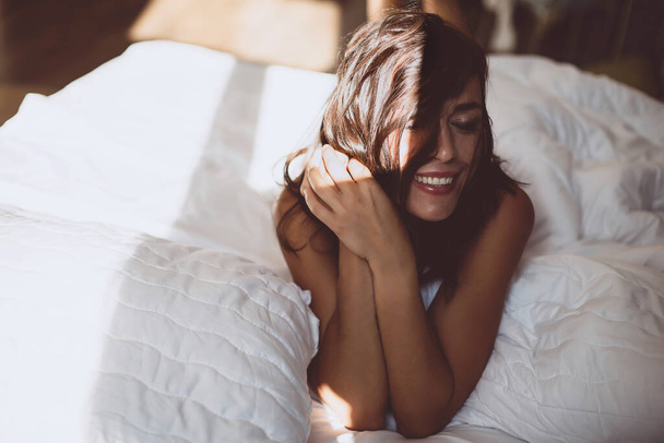 A beautiful young girl in the morning in a white bed after waking up, smiles at the new day and the sun from the window and covers her face with hair. Soft selective focus. Toned photo. - Photo, Image
