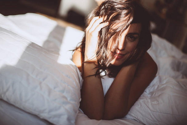 A beautiful young girl in the morning in a white bed after waking up, smiles at the new day and the sun from the window and covers her face with hair. Soft selective focus. Toned photo. - Photo, image