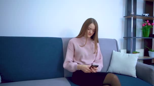 A young girl sits on a sofa is surfing in a smartphone. - Video