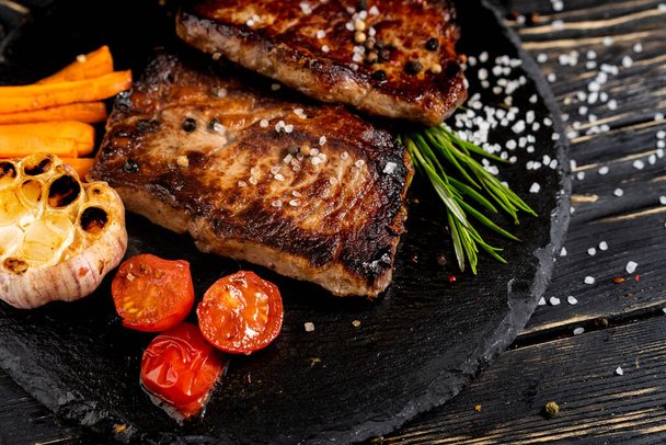 A juicy piece of fried meat with grilled cherry tomatoes garlic and carrot lies on a stone plate against a black wooden table. Degree of roasting well done. Food concept with copy space - Photo, Image