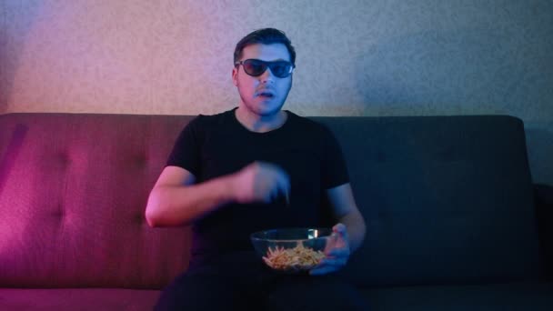 A man sits on a sofa in 3D glasses eating chips. - Záběry, video