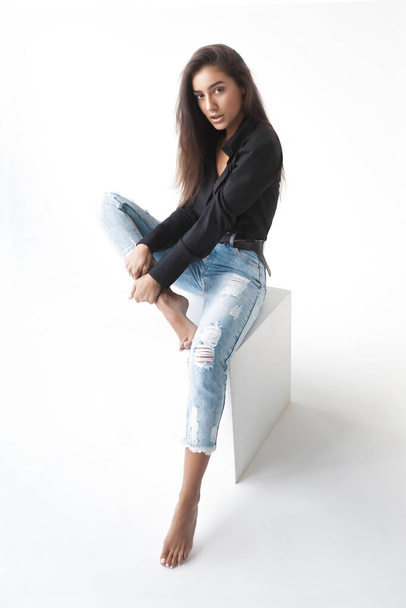Young cool hipster girl in black shirt and blue banana jeans girl sitting on white cube on white background. Fashionable modern style - Photo, Image