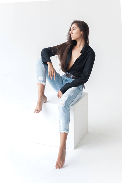 Young cool hipster girl in black shirt and blue banana jeans girl sitting on white cube on white background. Fashionable modern style - Zdjęcie, obraz
