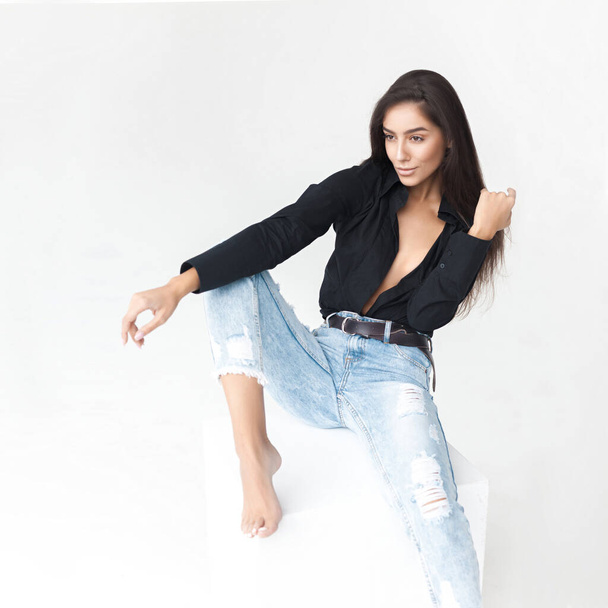 Young cool hipster girl in black shirt and blue banana jeans girl sitting on white cube on white background. Fashionable modern style - Foto, Imagem