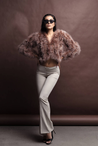 Young elegant girl posing in fluffy pink cape with artificial fur and beige flare trousers on brown background. Fashion retro style - Фото, зображення