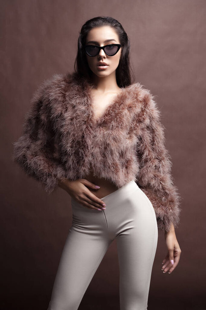 Young elegant girl posing in fluffy pink cape with artificial fur and beige flare trousers on brown background. Fashion retro style - Fotografie, Obrázek