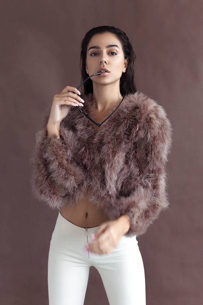 Young elegant girl posing in fluffy pink cape with artificial fur and beige flare trousers on brown background. Fashion retro style - Fotó, kép
