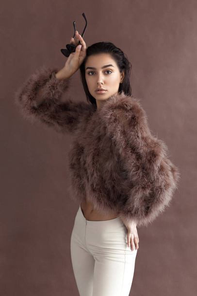 Young elegant girl posing in fluffy pink cape with artificial fur and beige flare trousers on brown background. Fashion retro style - Foto, afbeelding