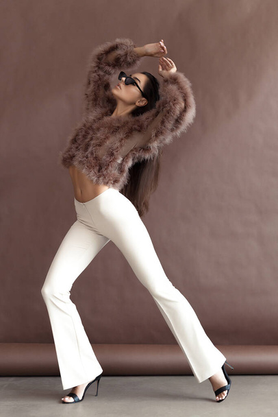 Young elegant girl posing in fluffy pink cape with artificial fur and beige flare trousers on brown background. Fashion retro style - Foto, immagini