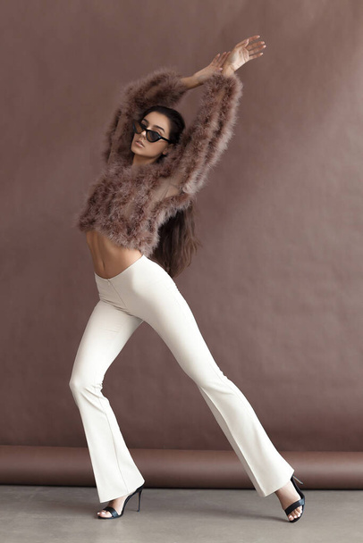 Young elegant girl posing in fluffy pink cape with artificial fur and beige flare trousers on brown background. Fashion retro style - 写真・画像