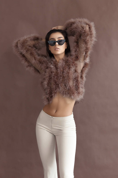 Young elegant girl posing in fluffy pink cape with artificial fur and beige flare trousers on brown background. Fashion retro style - Foto, Imagem