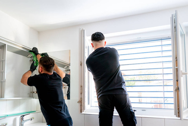 Una vista de dos limpiadores profesionales en un baño moderno limpiando los gabinetes y el espejo y persianas
 - Foto, imagen
