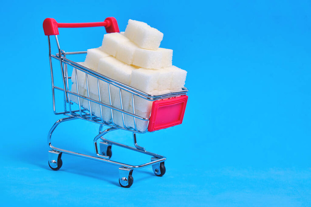 Shopping cart is filled with refined sugar cubes on a blue background - Photo, image