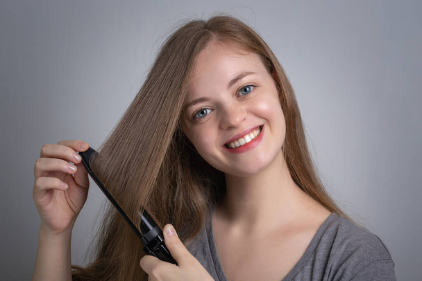 Joven mujer caucásica chica está alisando su pelo largo usando plancha de pelo eléctrico alisador con cara sonriente feliz
 - Foto, Imagen