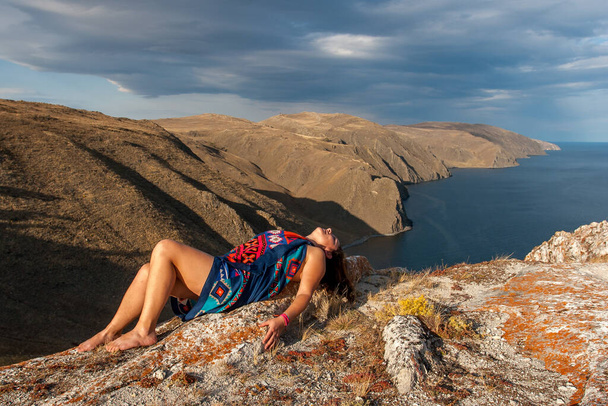 The girl lies on a rock above the lake. The model is wrapped in a scarf. In the background is a shore with beautiful cliffs. There are clouds in the sky. - Photo, Image