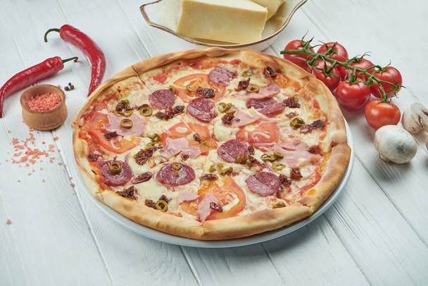 Classic italian pizza with mozzarella, tomatoes, olives, mushrooms, salami and ham on a white wooden background in a composition with ingredients. - Foto, immagini