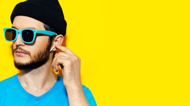 Studio πορτρέτο του νεαρού hipster άνθρωπος σε μπλε πουκάμισο, φορώντας κυανό γυαλιά ηλίου και μαύρο καπέλο σκούφο, χρησιμοποιώντας ασύρματα ακουστικά σε φόντο κίτρινο χρώμα με χώρο αντίγραφο. - Φωτογραφία, εικόνα