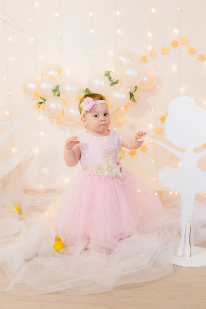 Cute little girl in a pink princess dress with a flower on her hair plays with wooden ballerinas among the golden stars on the background of a large white wooden moon and balloons. Children's decor.  - Fotografie, Obrázek