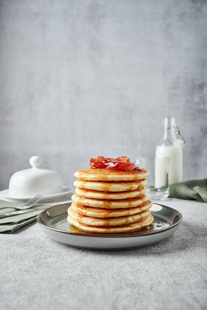 American-style pancakes with apple jam in muted green plate on grey background. Bottle of milk, butter-dish and green napkin on the background. Fork and knife on another napkin near the plate - Foto, Imagem