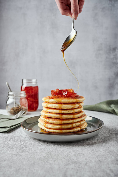 American-style pancakes with apple jam in muted green plate on grey background. A jar of jam, tea and green napkin on the background. Fork on another napkin near the plate. Hand pours honey on top of pancakes - Foto, immagini