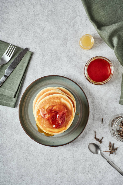 American-style pancakes with apple jam in muted green plate on grey background. Fork and knife on a napkin, jars with honey, tea spoon and some tea on the table - Фото, изображение