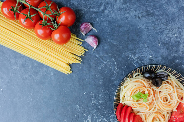Spaghetti made from durum wheat and ripe cherry tomatoes on a branch, garlic and spices. Fresh greens on a dark graphite background. Products for making delicious pasta. The view from the top. - Foto, Bild