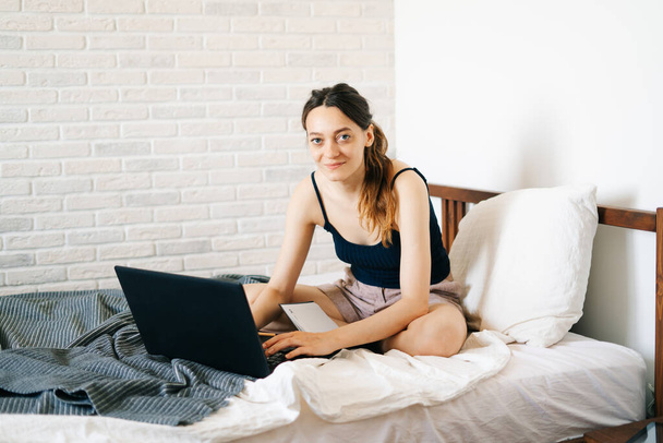 Μια νεαρή γυναίκα, χρησιμοποιώντας ένα φορητό υπολογιστή, επικοινωνεί μέσω του Διαδικτύου με έναν πελάτη στο κρεβάτι της, ενώ ο ιός είναι σε καραντίνα. Άνετος χώρος εργασίας γραφείου, Απομακρυσμένη εργασία, e-learning έννοια - Φωτογραφία, εικόνα