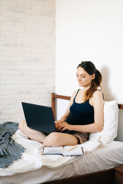 A young woman, using a laptop, communicates over the Internet with a client in her bed, while the virus is quarantined.Cozy office workplace, Remote work, e-learning concept - Фото, изображение